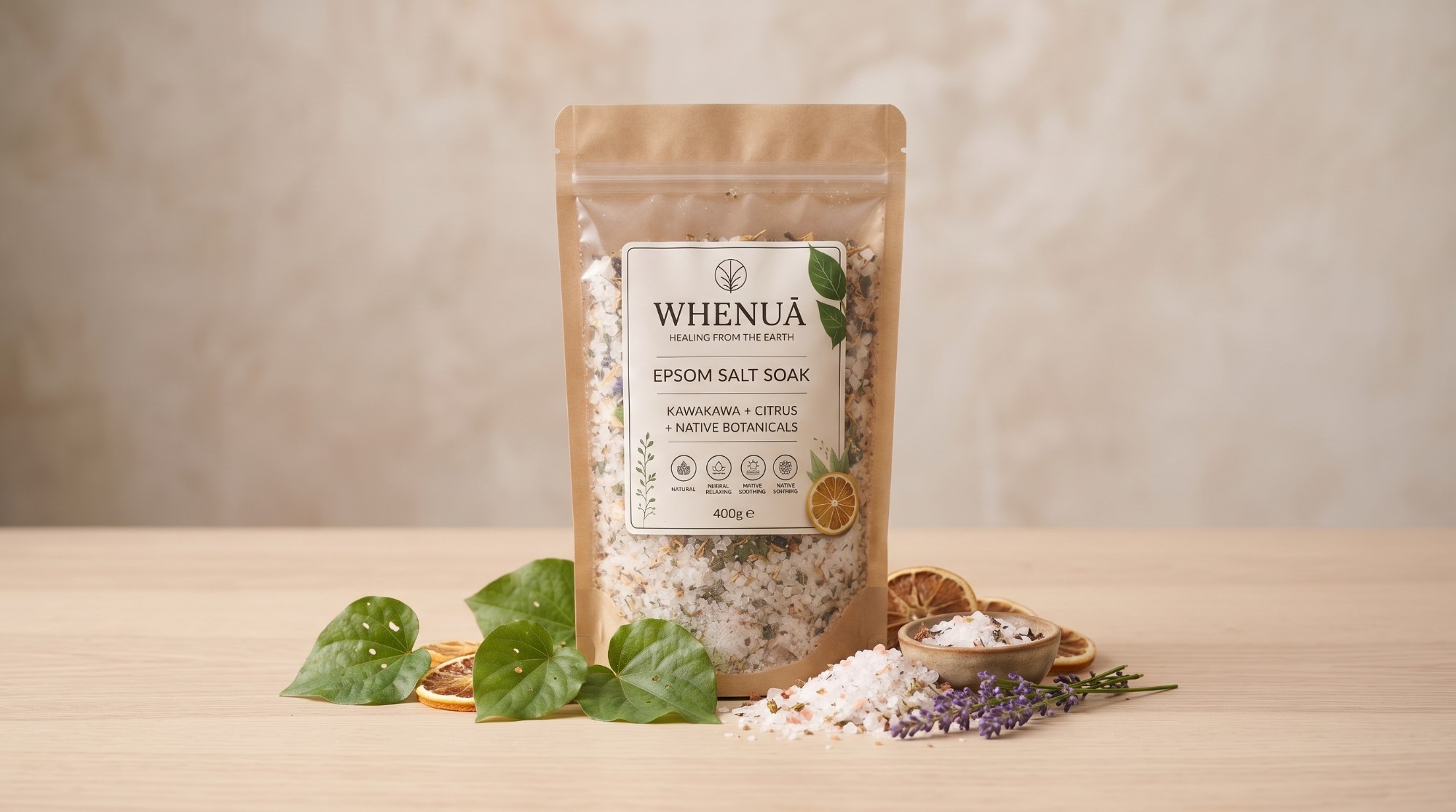 The Whenuā pouch standing on pale wood with kawakawa leaves, dried citrus and lavender arranged around it.